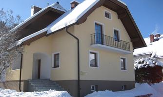 A yellow house with a roof and a balcony, surrounded by snow. The sky is clear and blue.