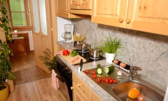 A cozy kitchen with wooden furniture and plants. Fresh vegetables are on the countertop next to a stove and a sink.