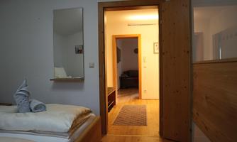 An inviting room with two beds and a wooden floor. The door leads into a bright hallway that connects to other rooms.