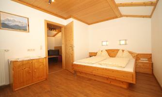 A cozy bedroom with a wooden double bed and a warm light entrance. A landscape painting hangs on the wall, and a wooden wardrobe complements the decor.