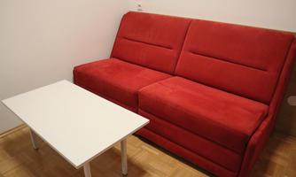 A red sofa stands against a wall next to a small white table. The room has a simple and modern look.