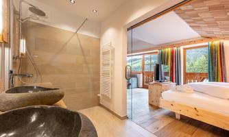 A modern bathroom with two sinks and a glass shower. In the background, a cozy bedroom with colorful curtains is visible.