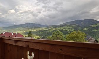 A scenic view with hills and mountains under a cloudy sky. In the foreground, a wooden railing is visible.