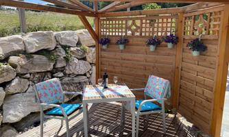 An inviting seating area with a table and two chairs under a wooden overhang. Flower plants hang on the wall, and a bottle of wine sits on the table.