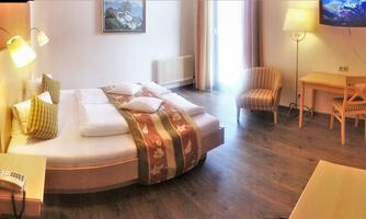 A cozy hotel room with a double bed, a wooden chair, and a table. The walls are light, and the room has large windows with curtains.