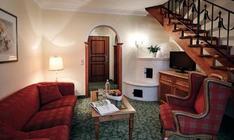 A cozy living room with a red sofa and an armchair. A wooden staircase leads to an upper area, and a table stands in the center of the room.