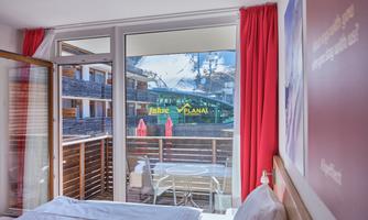 A cozy room with a balcony that offers a beautiful view of the mountains. Through the windows, one can see clear skies and bright colors.