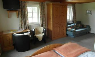 A cozy room with a double bed, a wardrobe, and a sofa. Large windows provide plenty of daylight.