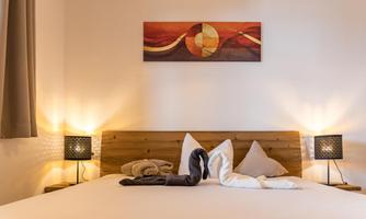 A cozy bedroom with a large bed and modern nightstand lamps. Above the bed hangs an abstract artwork in warm colors.