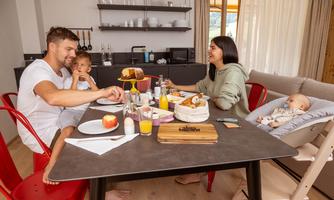 A family is sitting at the table and enjoying breakfast together. Various foods and drinks are on the table, while a baby is sitting in a high chair.