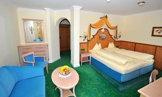 A cozy hotel room with a double bed, sofa, and a small table. The walls are painted light and the floor is covered with green carpet.