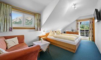 A cozy bedroom with two beds and a sofa. The room has windows with a view of the mountains and a balcony.
