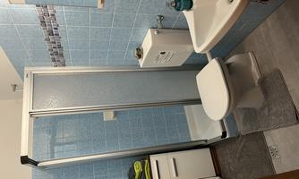 A small bathroom with blue tiles and a shower. There is a toilet, a sink, and storage space.