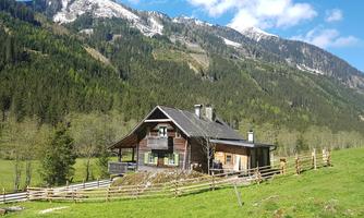 A cozy wooden house in a green mountain landscape. In the background, snow-capped mountains and a blue sky can be seen.