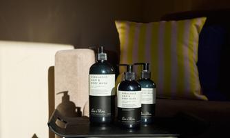 Three bottles of body care products on a tray. In the background, an elegant sofa and a decorative pillow can be seen.