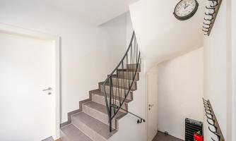 A clear hallway with a curved staircase and a wall coat rack. A black wall clock is hanging on the wall.