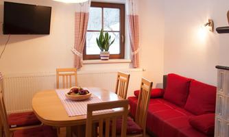A cozy living space with a round dining table and red chairs. In the background, there is a red sofa and a television on the wall.