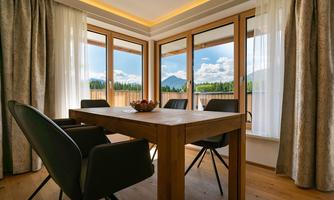 A bright apartment with a wooden table and modern chairs. Large windows offer a view of a mountain landscape and the blue sky.