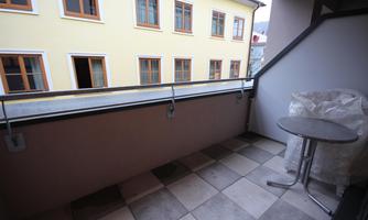 A balcony with a small table and a chair. In the background, there is a yellow building with large windows.