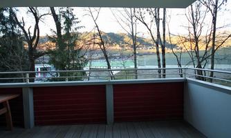 A balcony with a wooden floor and a red railing. Trees and gentle hills can be seen in the background.