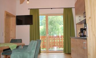 A modern kitchen with a dining table and comfortable chairs. Large windows with green curtains offer a view of the greenery.