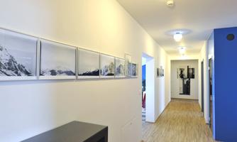 A bright hallway with wooden flooring and white walls. Framed black-and-white photos of mountains hang on the wall.