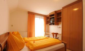 A bright bedroom with two beds and orange bedding. There is a desk, a television, and large windows with curtains.