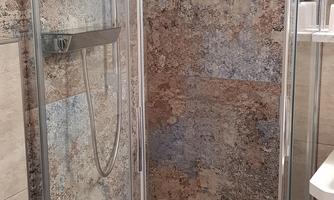 A modern shower cubicle with glass walls and an appealing mosaic pattern. The floor is equipped with a blue bath mat.