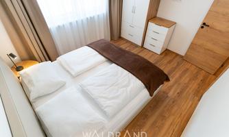 A modern bedroom with a double bed and a wooden floor. Next to the bed is a nightstand and a dresser.