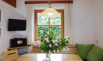 A bright dining area with a wooden table and a vase full of white flowers. The windows offer a view of a green garden, and there are cozy cushions.