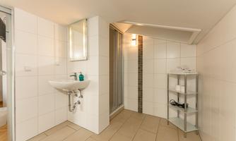 A modern bathroom with white tiles and a sink. On the right side, there is a shelf with towels and personal care products.
