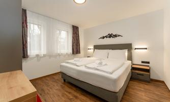 A modern bedroom with a double bed and wooden flooring. Large windows provide plenty of daylight.