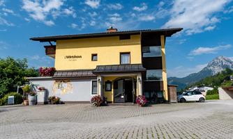 A pretty yellow house with a modern design, surrounded by plants and flowers. In the background, there are mountains and a blue sky.