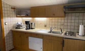 A small kitchen with wooden cabinets and a bright workspace. There is a coffee machine, a kettle, and a stove.