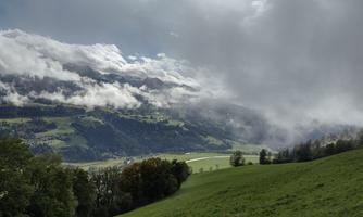 A picturesque landscape with rolling hills and dense clouds. The green meadow stretches to the horizon, where the mountains are visible.