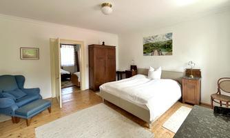 A cozy bedroom with a double bed and a blue armchair. The walls are light, and there is a picture of a landscape.