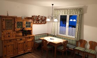 A cozy dining area with wooden furniture and a windowsill. Next to a table with chairs, there is a small seating area and a display cabinet.