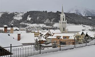 A picturesque winter landscape with snow-covered houses and a church in the foreground. In the background, snow-covered mountains rise under a gray sky.