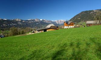 An idyllic landscape with green meadows and traditional farmhouses. In the background, majestic mountains rise under a radiant blue sky.