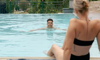 A man is swimming in the pool while a woman is sitting at the edge. The scene is relaxed and sunny.