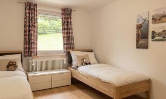A bright bedroom with two beds and wooden furniture. The room has large windows overlooking nature and decorative pictures on the wall.