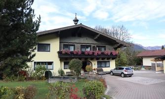 A charming house with blooming balconies and a green environment. In the foreground, there is a car and well-maintained gardens.