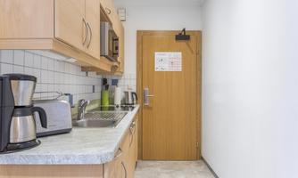 A small, modern kitchen with wooden cabinets and a stainless steel sink. On the countertop are a coffee machine and a toaster.