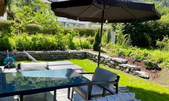 A cozy garden with a table and chairs under a sun umbrella. In the background, there are green plants and a well-kept landscape.