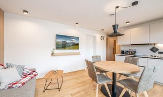 A modern living room with a round dining table and comfortable chairs. An artwork and a clock hang on the wall, while a cozy couch invites relaxation.