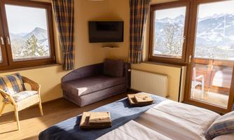 A cozy bedroom with two beds, a sofa, and large windows that offer a view of the mountains. The decor is bright and inviting.