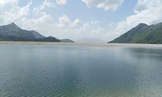 A peaceful lake surrounded by mountains and gentle hills. The sky is partly cloudy and reflects in the water.