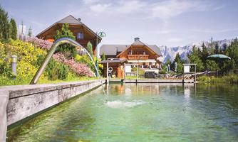 A beautiful lake with clear water and surrounded by flowers. In the background, a cozy wooden house and the mountains can be seen.