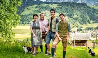 A group of four people stands in a meadow with a picturesque mountain landscape in the background. They are wearing traditional costumes and smiling at the camera.