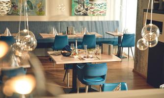 A modern restaurant with blue chairs and elegant tables. The walls are decorated with colorful paintings, and the lighting is soft.
